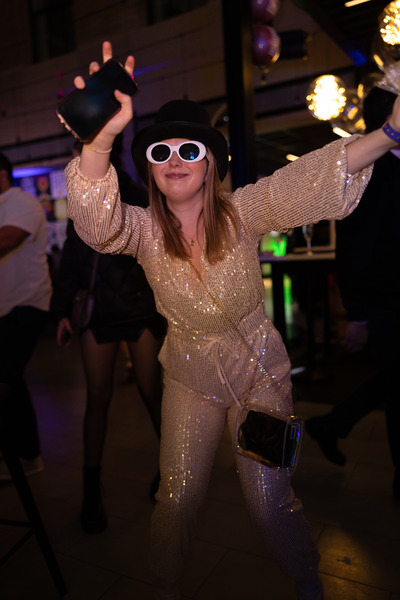 A girl in a gold sparkly outfit and white sunglasses dances at High Street Place. 